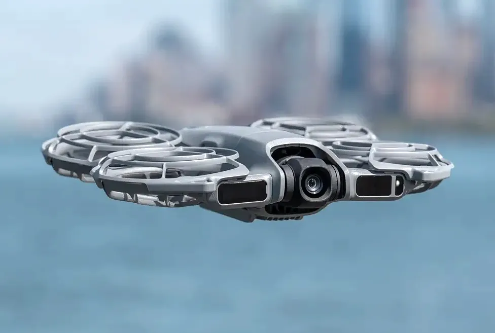 Close-up of a gray quadcopter drone with propeller guards flying outdoors over blurred blue water, with a city skyline in the background—perfect for showcasing advanced tech like Teams direct routing SMS solution integrations.